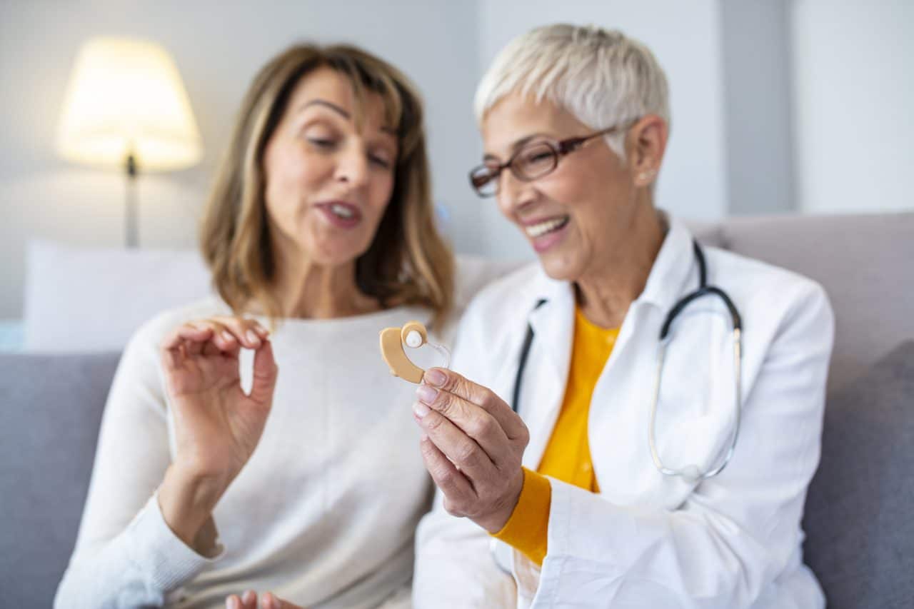 Mature female doctor hearing specialist in her office trying hearing aid equipment to a patient elderly senior woman. The doctor laryngologist explains to senior woman how to wear a hearing aid